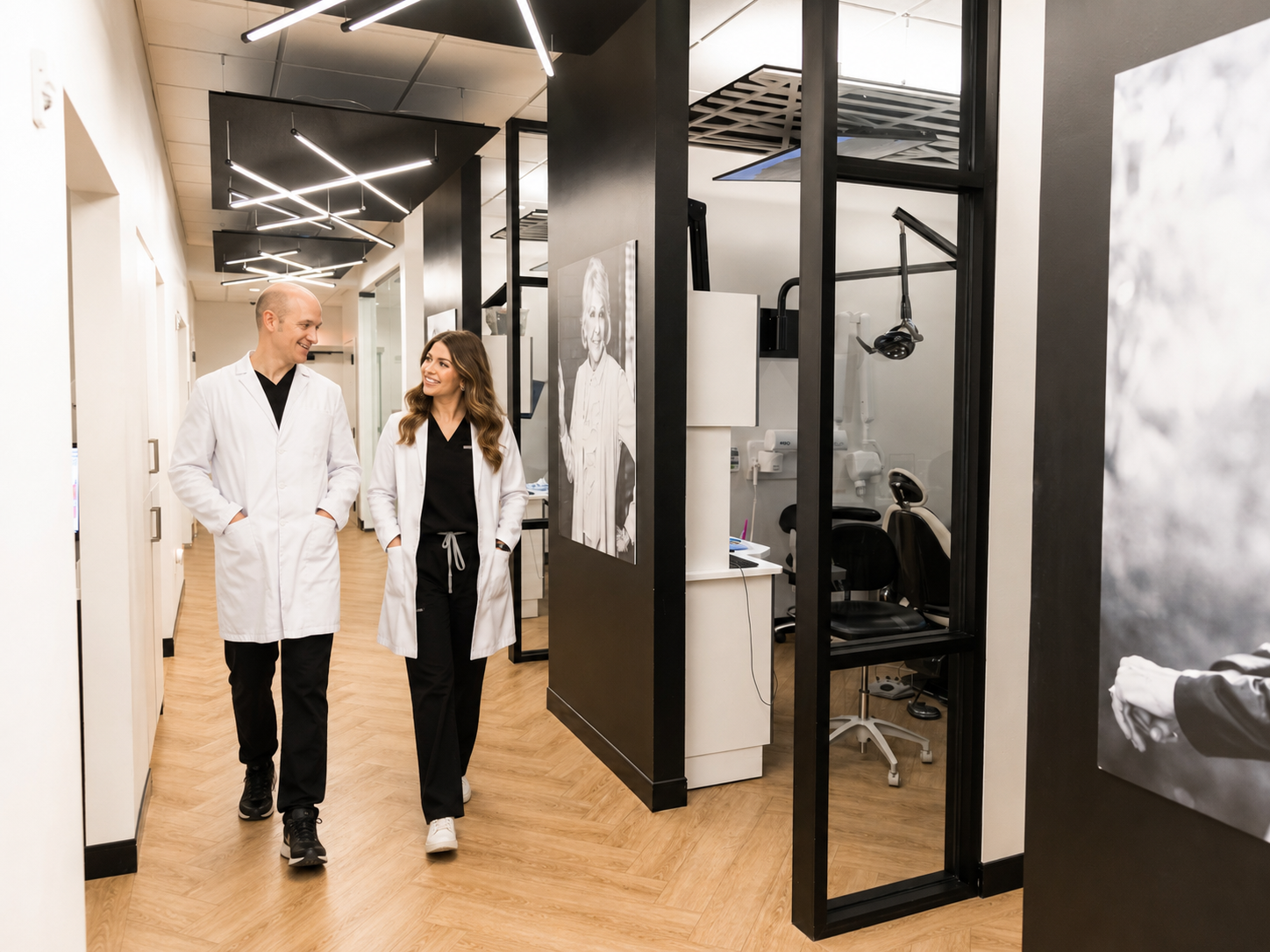 Two dental professionals in white coats walking through a modern dental office hallway past open treatment rooms